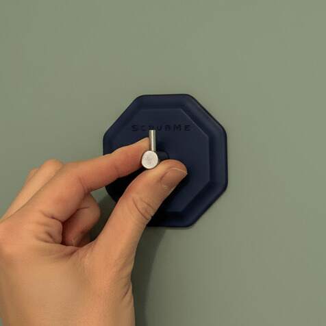 Pressing the ScrubMe HookMe Breathtaking Blue adhesive hook onto a soft green wall to stick it firmly in place.