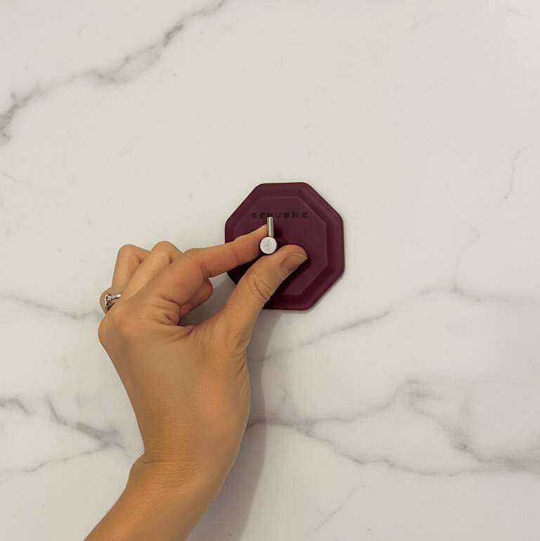 Hand installing the ScrubMe HookMe Passionate Purple adhesive hook onto a smooth marble bathroom wall.