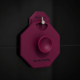 ScrubMeBody Passionate Purple silicone body scrubber hanging on a chrome hook against sleek black bathroom tiles.