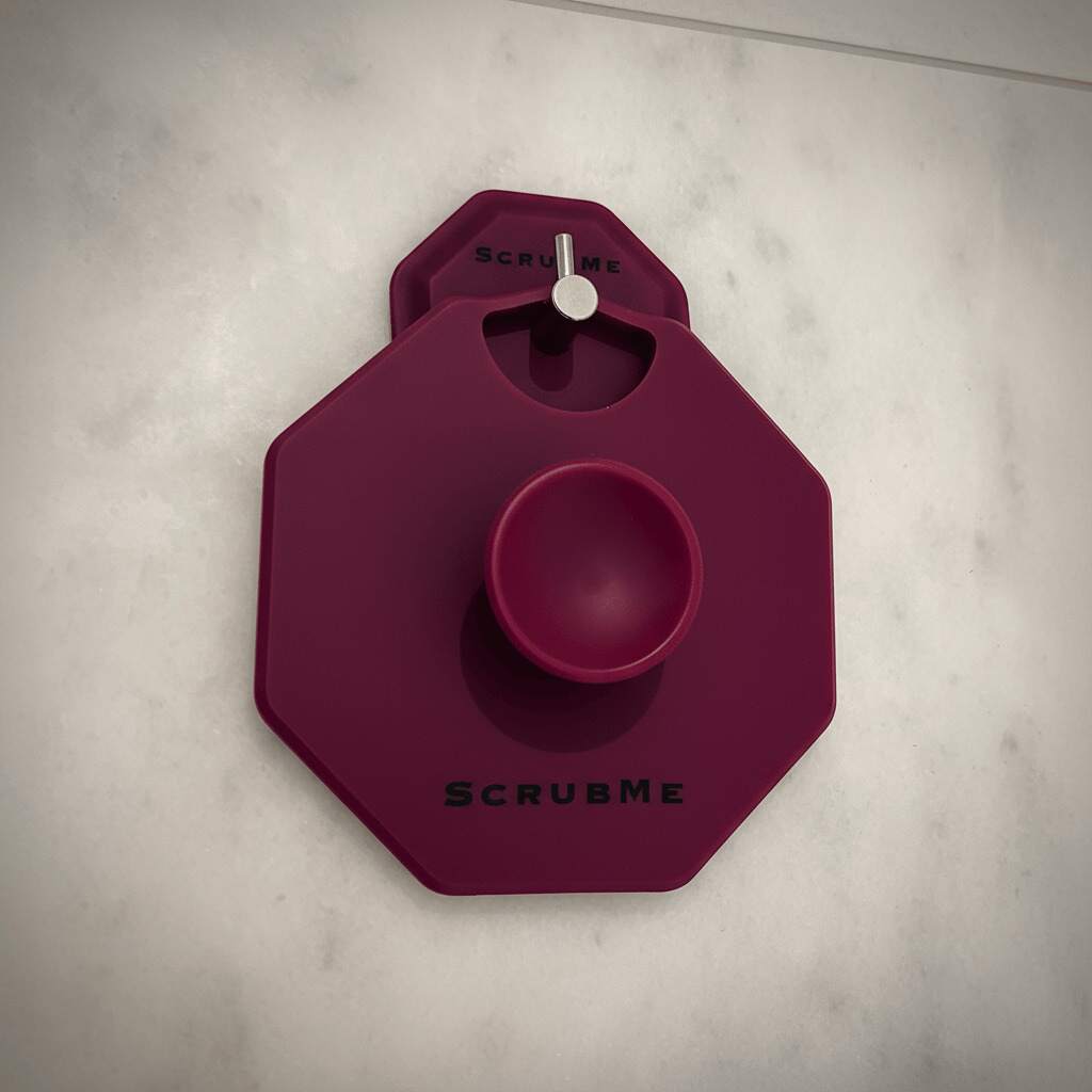 ScrubMeBody Passionate Purple silicone body scrubber hanging neatly on a wall hook against light marble shower tiles.