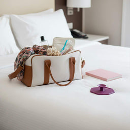 ScrubMeBody Passionate Purple body scrubber on a hotel bed beside an open overnight bag, scarf and toiletries.