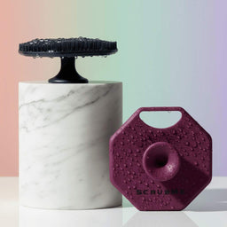 Hero Image ScrubMeFace purple silicone face scrubber and blue bristle stand displayed on marble plinths with pastel background.