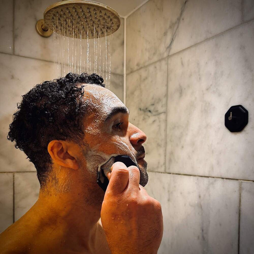Man in a marble shower using the ScrubMeFace Breathtaking Blue face scrubber to massage shaving foam into his skin.
