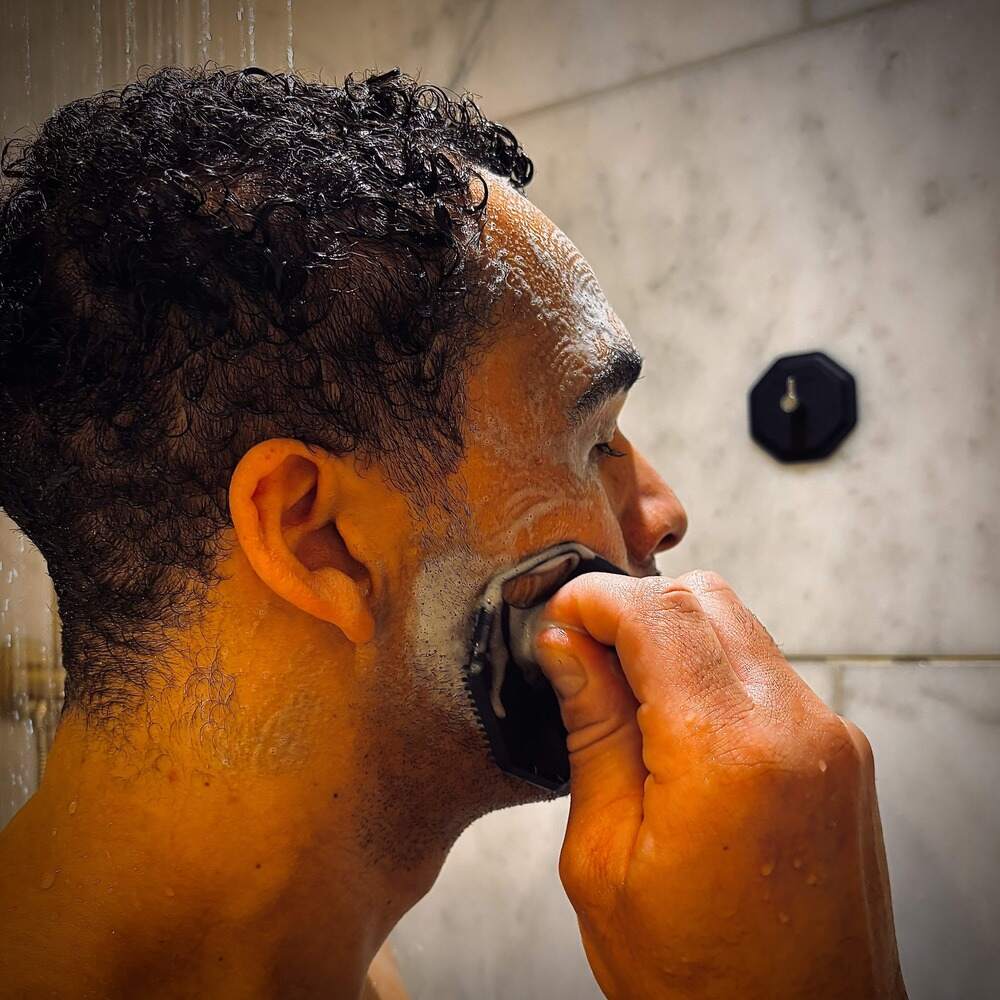 Side view of a man shaving in the shower while massaging his face with the ScrubMeFace Breathtaking Blue silicone scrubber.
