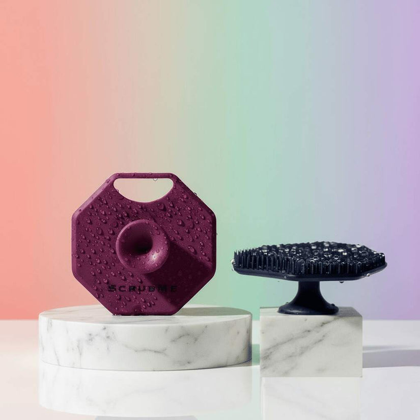 ScrubMeFace silicone face scrubbers in Passionate Purple and Breathtaking Blue on marble stands with water droplets against a pastel background