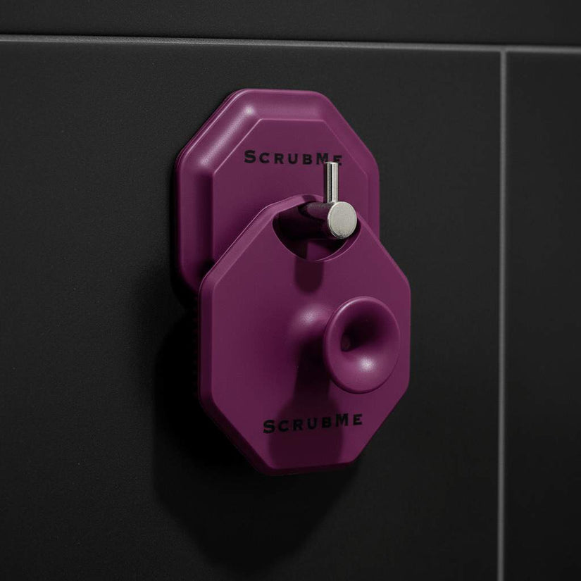 ScrubMeFace Passionate Purple silicone face scrubber hanging on a wall hook against black shower tiles.