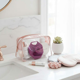 ScrubMeFace Passionate Purple silicone face scrubber in a clear vanity travel bag with skincare and sleep accessories on a bathroom counter.