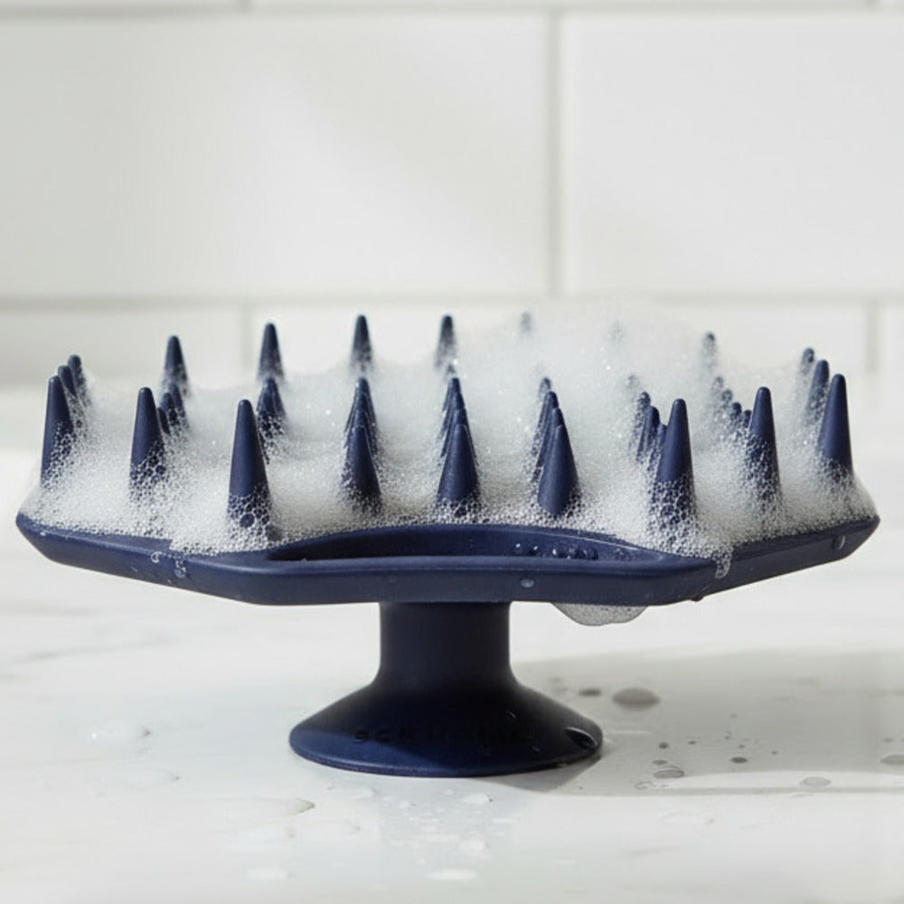 ScrubMeScalp Breathtaking Blue silicone scalp scrubber standing on its suction base with foaming bristles on a white tiled bathroom surface