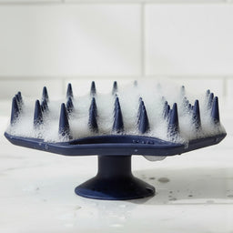 ScrubMeScalp Breathtaking Blue silicone scalp scrubber standing on its suction base with foaming bristles on a white tiled bathroom surface