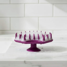 ScrubMeScalp Passionate Purple silicone scalp scrubber standing on its suction base with foaming bristles on a white tiled bathroom sink
