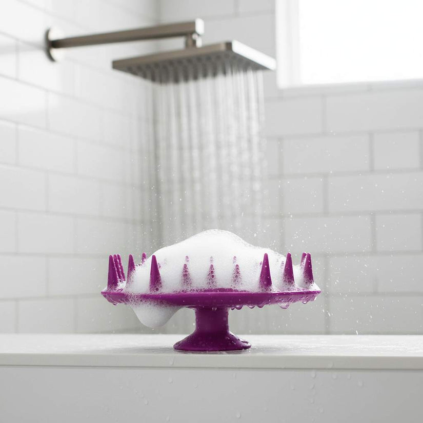 ScrubMeScalp Passionate Purple scalp scrubber covered in foam on a shower ledge with rainfall shower running against white subway tiles