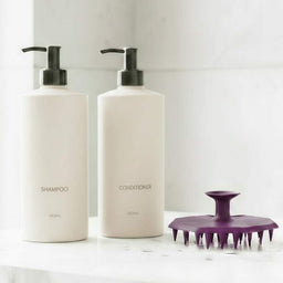 ScrubMeScalp Passionate Purple scalp scrubber standing on a bathroom ledge next to minimalist white shampoo and conditioner bottles