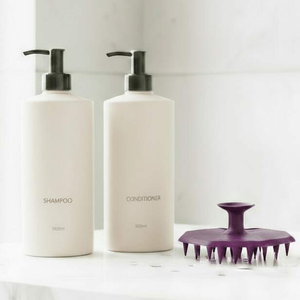 ScrubMeScalp Passionate Purple scalp scrubber standing on a bathroom ledge next to minimalist white shampoo and conditioner bottles